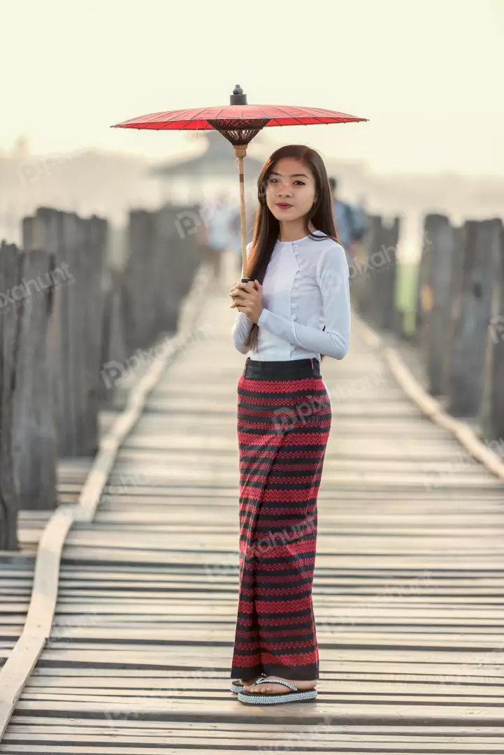 Free Premium Stock Photos A beautiful Burmese woman in traditional dress and girl standing on a wooden bridge, holding a red umbrella