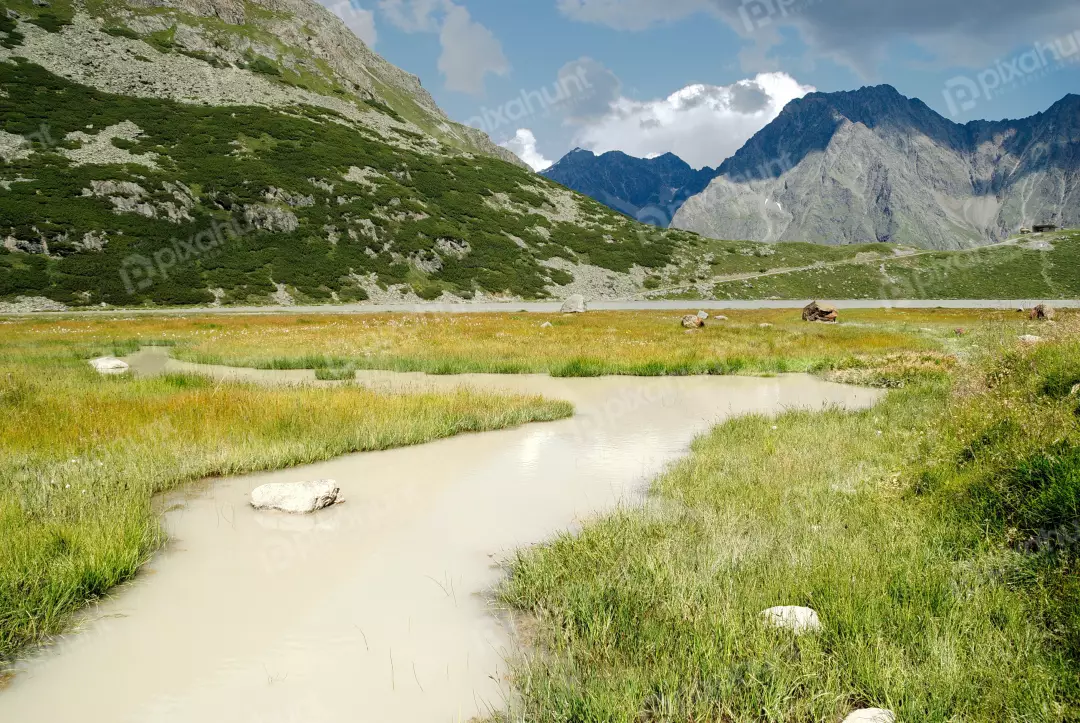Serene Alpine Meadow with Winding Stream and Majestic Mountains Under Cloudy Sky