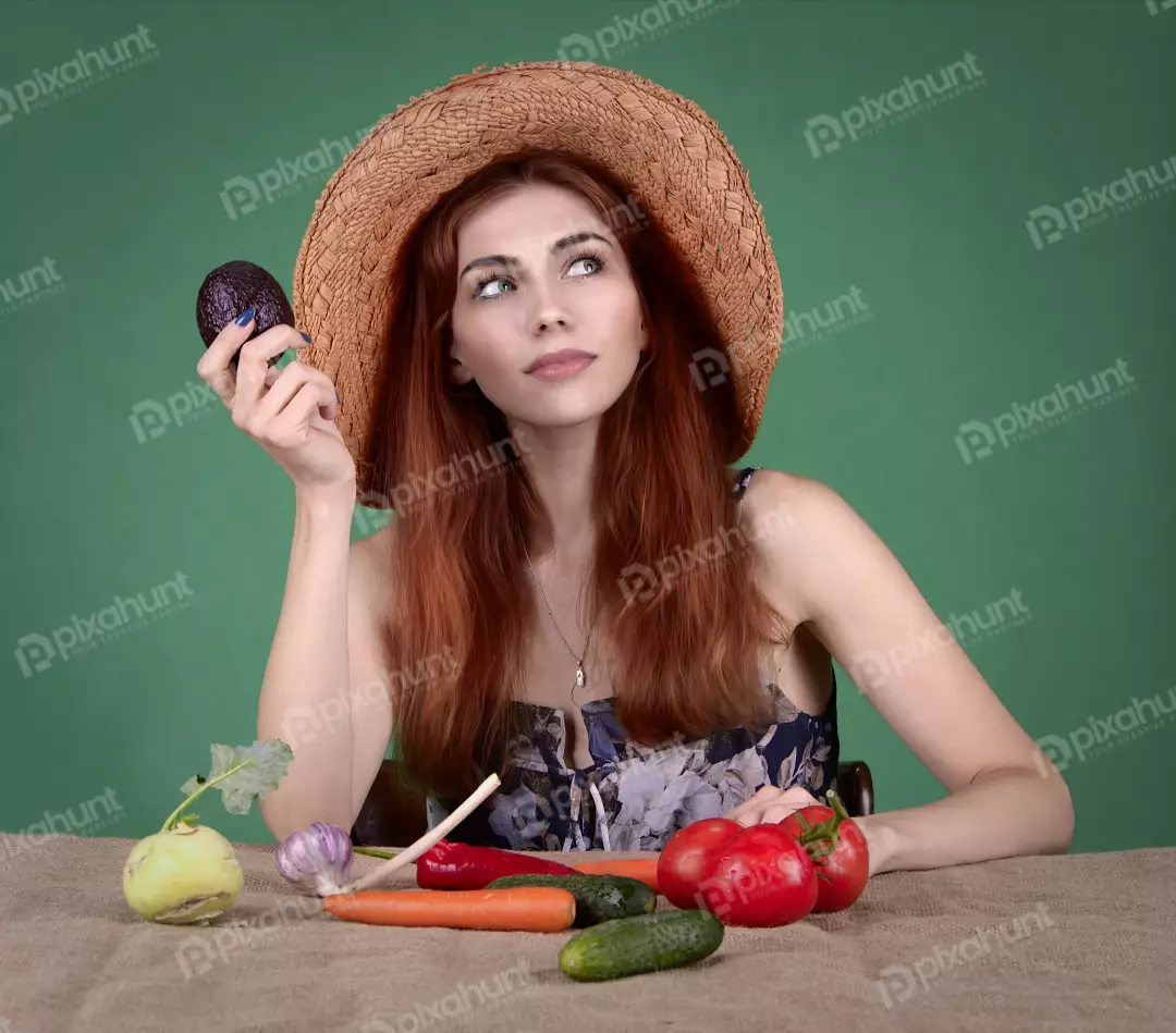 Thoughtful Young Woman in Straw Hat Holding Avocado with Fresh Vegetables