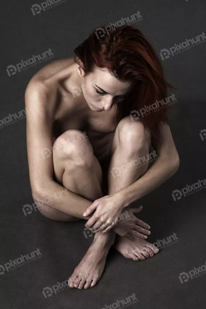 Red-haired woman sitting nude in a vulnerable, contemplative pose on a dark background