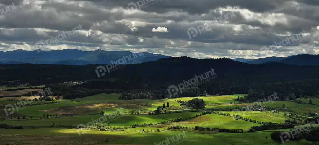 Dramatic Sunbeams Illuminate Verdant Fields and Layered Mountains Under an Overcast Sky