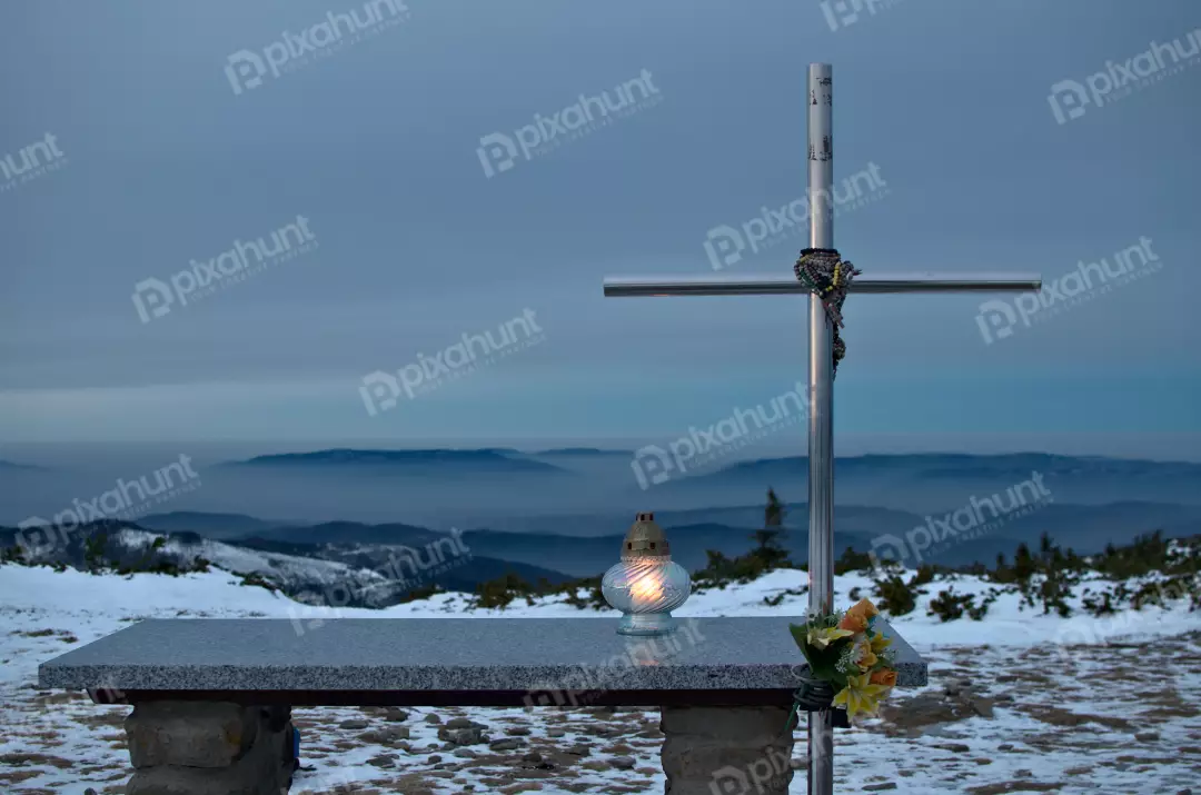 Glowing Candle by Metal Cross on Snowy Mountain Peak at Twilight