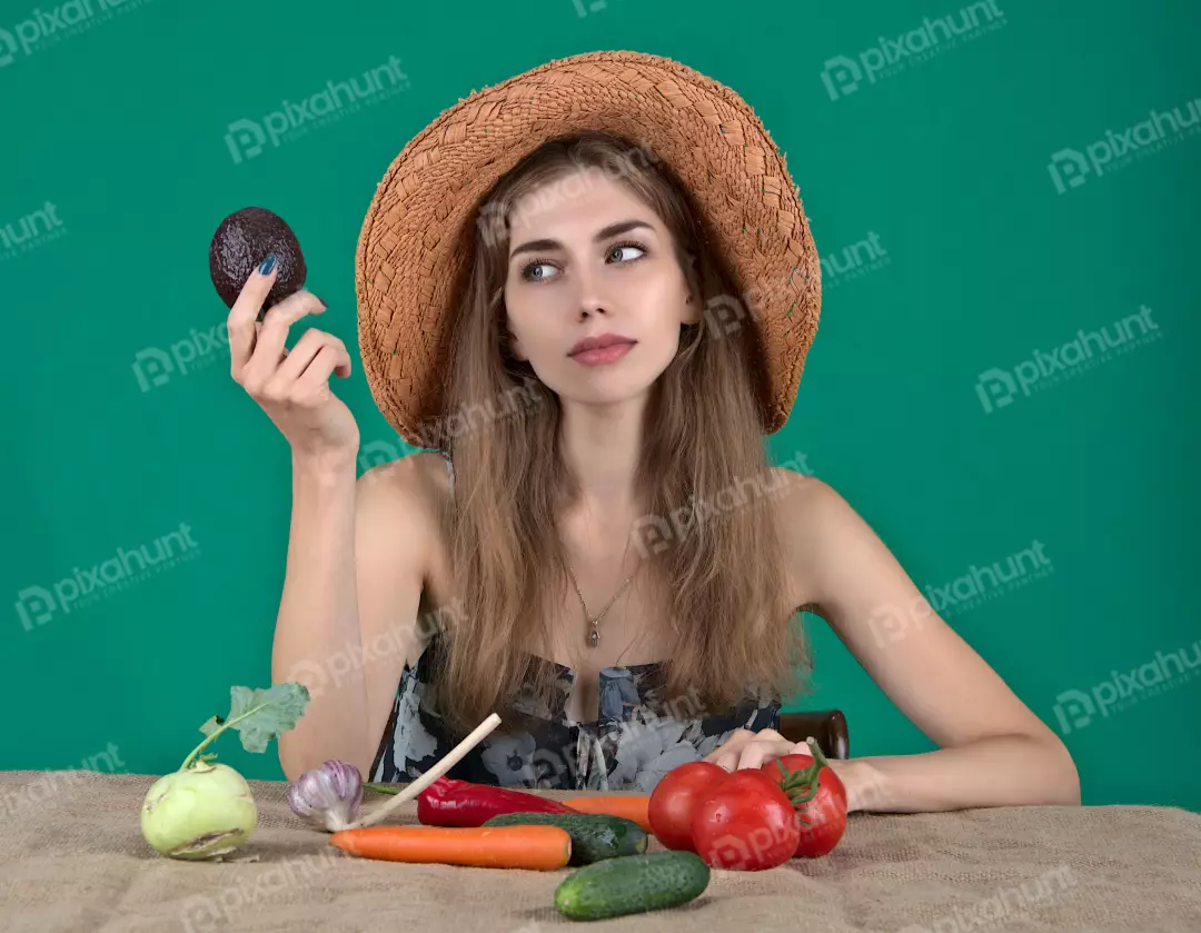 Thoughtful Woman in Straw Hat Holding Avocado with Garden Vegetables