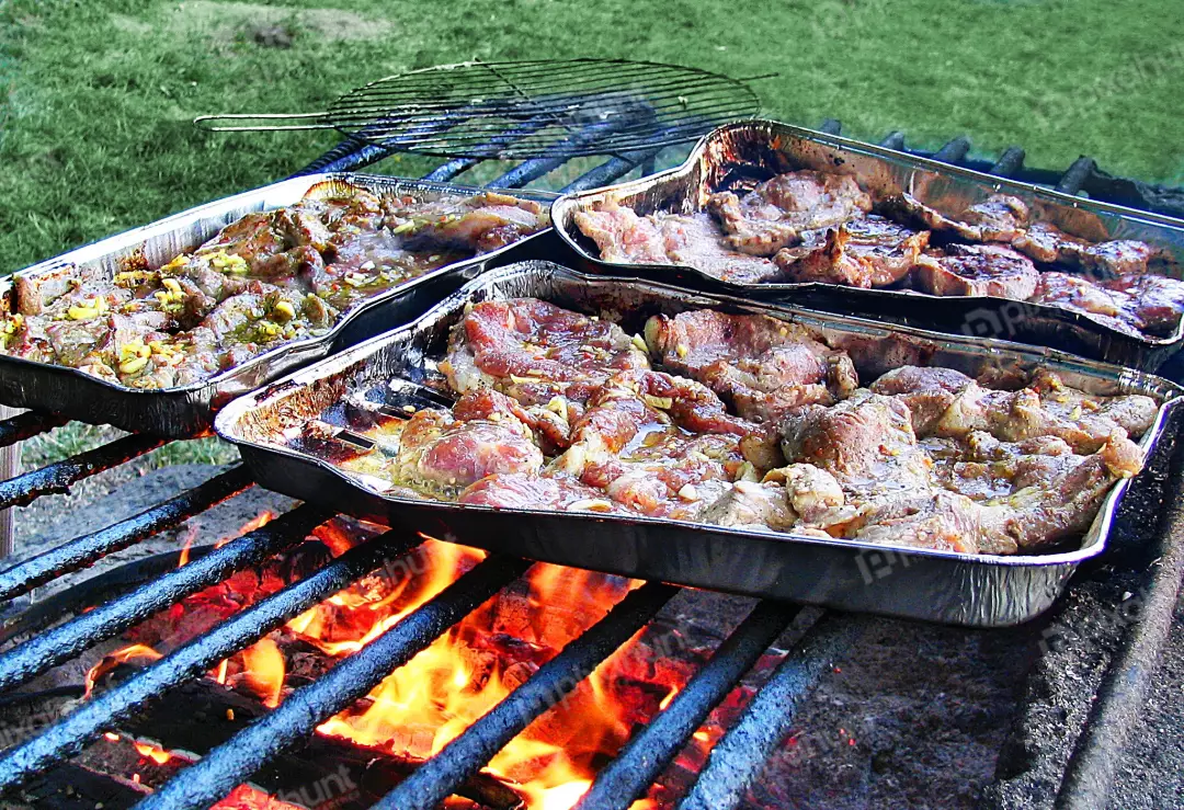 Marinated meat sizzling on outdoor barbecue grill in foil pans over hot coals
