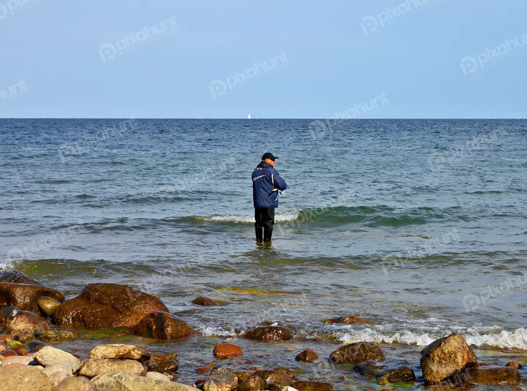 Man Fishing in Serene Sea with Rocky Shore and Distant Sailboat