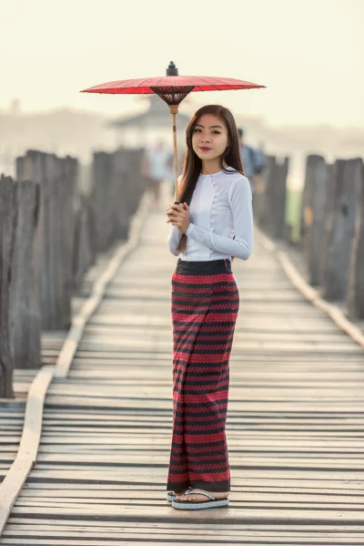 A beautiful Burmese woman in traditional dress and girl standing on a wooden bridge, holding a red umbrella