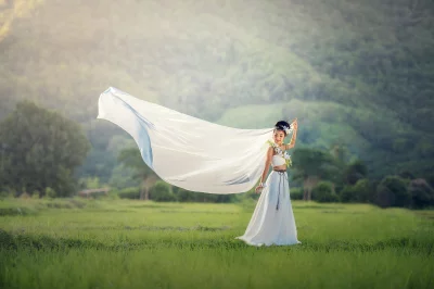 A beautiful young girl wearing a white dress and standing in a field of tall grass and the wind is blowing her hair and dress