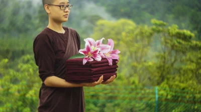 Monk Holding A Stack Of Red Cloths With Flower