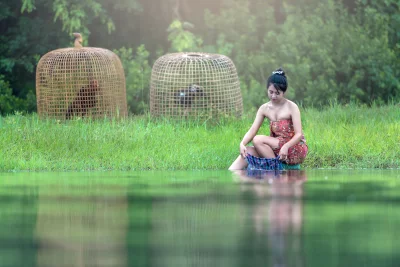 A young girl in a traditional Thai outfit sitting on the edge of a river and washing clothes in the water