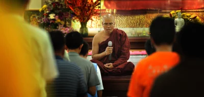 Buddhist monk sitting on a raised platform, with a microphone in his hand