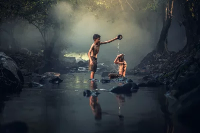 Two boys who are bathing in a river and standing in the water and the one on the left is pouring water over the head of the boy on the right