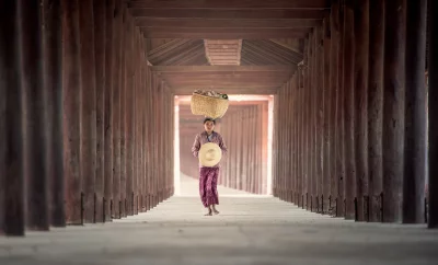 A woman who is walking away from the camera and carrying a large basket on her head and is wearing a traditional Burmese longyi