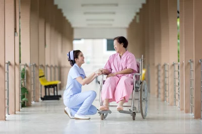 A nurse kneeling next to a patient in a wheelchair and nurse is smiling and holding the patient's hand