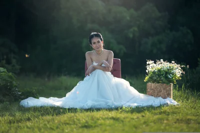 The bride is sitting on a red chair in the middle of a field and wearing a white wedding dress and has her hair in a bun