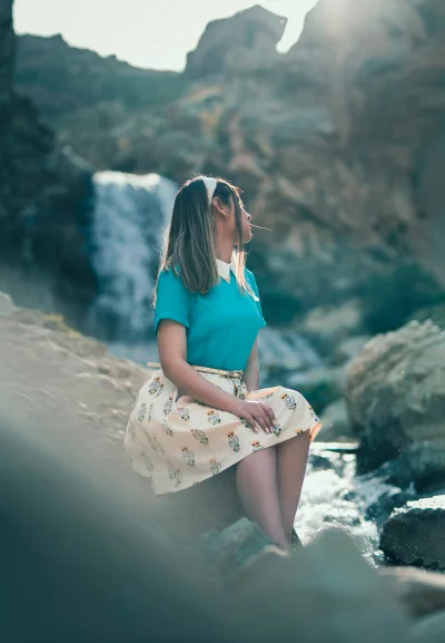 Young Woman in Teal Shirt and Patterned Skirt Relaxing by a Natural Waterfall