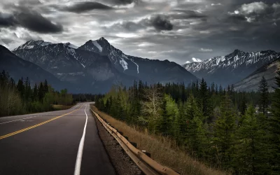 Scenic Winding Road Through Pine Forest with Majestic Snow-Capped Mountains Under a Dramatic Cloudy Sky image