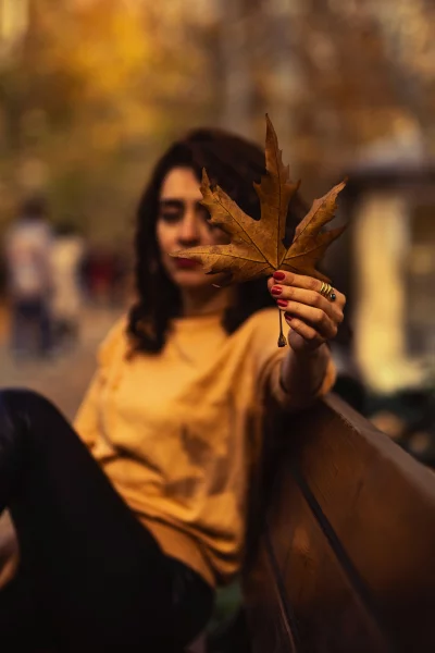 Blurred woman in a cozy yellow sweater welcoming the fall season and holding a crisp maple leaf in the foreground