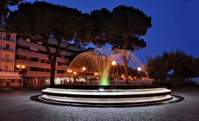 Illuminated Urban Fountain at Twilight with Pine Trees and Building image