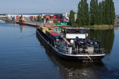 A large ship in a river ship is carrying a variety of containers and there are trees and buildings