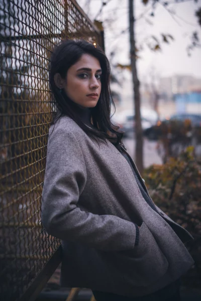 Pensive Young Woman in Beige Coat Leaning Against Rustic Metal Fence Outdoors