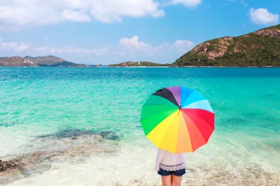 Person holding a vibrant rainbow umbrella on a tropical beach with clear blue ocean and islands image