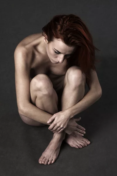 Red-haired woman sitting nude in a vulnerable, contemplative pose on a dark background image