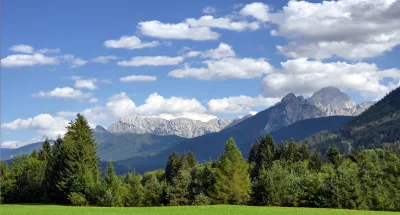 Majestic Alpine Landscape with Lush Green Meadow and Forested Mountains Under Cloudy Blue Sky image