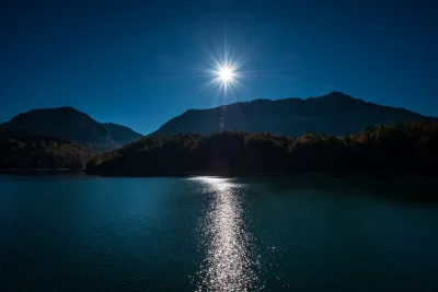 Radiant sunburst illuminates calm mountain lake with shimmering water reflection against dark forests image