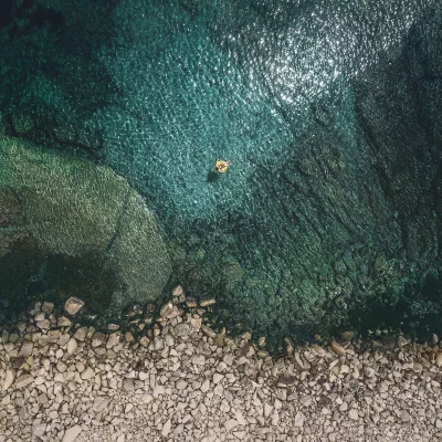 Aerial Drone View: Tranquil Turquoise Ocean with Person Floating on Yellow Inflatable by Rocky Beach image