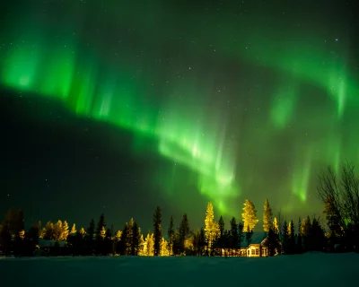 Vibrant Aurora Borealis over a tranquil winter landscape with illuminated forest and cabin image