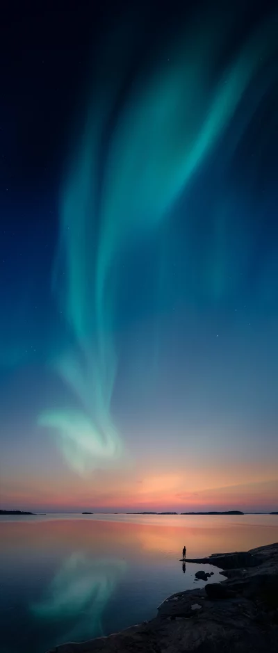 Person watches mesmerizing aurora borealis dance over serene water at dusk
