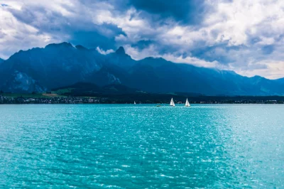 Serene Turquoise Lake with Distant Sailboats and Majestic Cloudy Mountains Landscape image