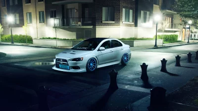 White Mitsubishi Lancer Evolution with Blue Rims Parked on Wet Urban Street at Night image