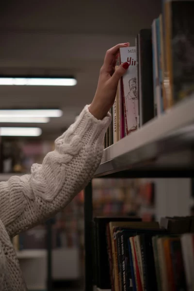 Hand reaching for book on a library shelf, cozy knitted sweater