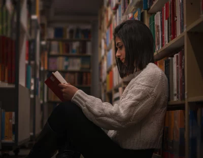 Young woman sitting between bookshelves reading a book in a library