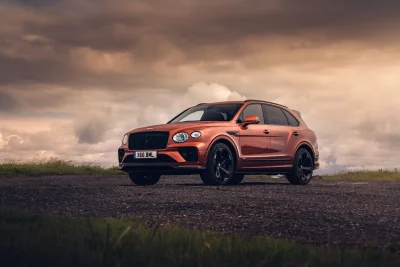 Luxury Orange Bentley Bentayga SUV on Gravel Road under Dramatic Sky Cinematic Wallpaper image