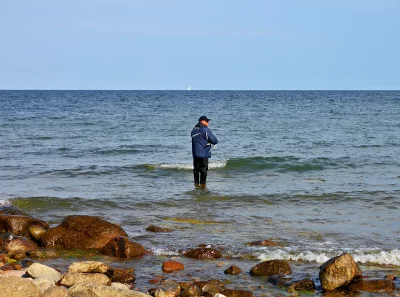 Man Fishing in Serene Sea with Rocky Shore and Distant Sailboat image